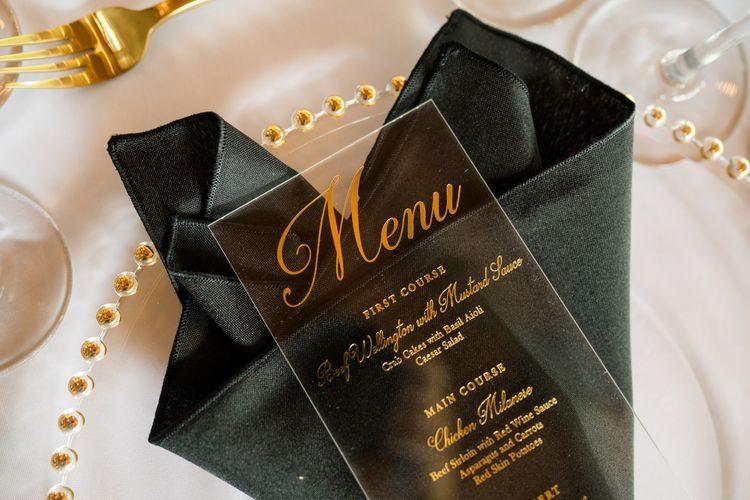 Acrylic menu with gold foil script resting on a black satin napkin with gold flatware